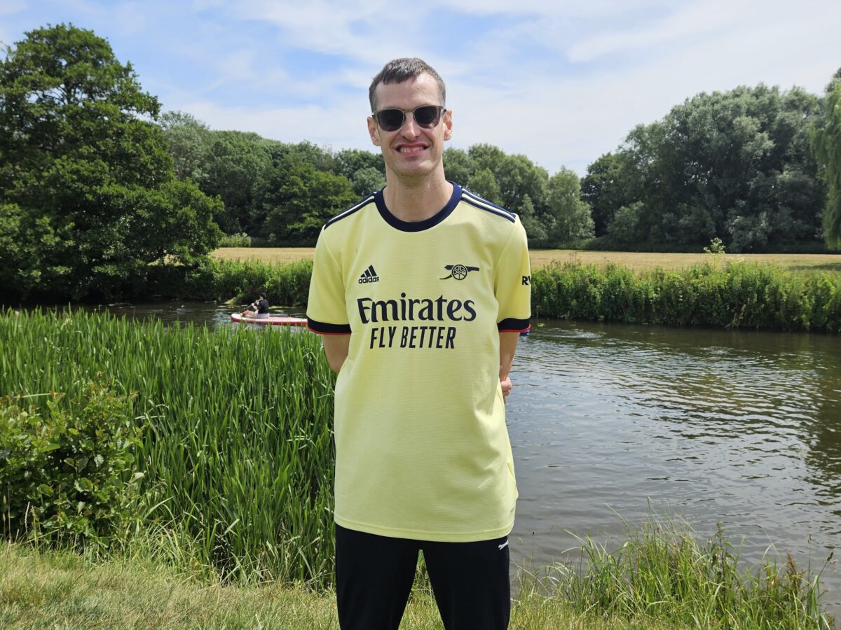 Mark smiling, standing in front of a river with a canoe passing.