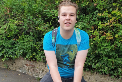 A blonde teenager wearing a blue tshirt with green graphic and a green backpack. He's posing on a statue of a hippopotamus.