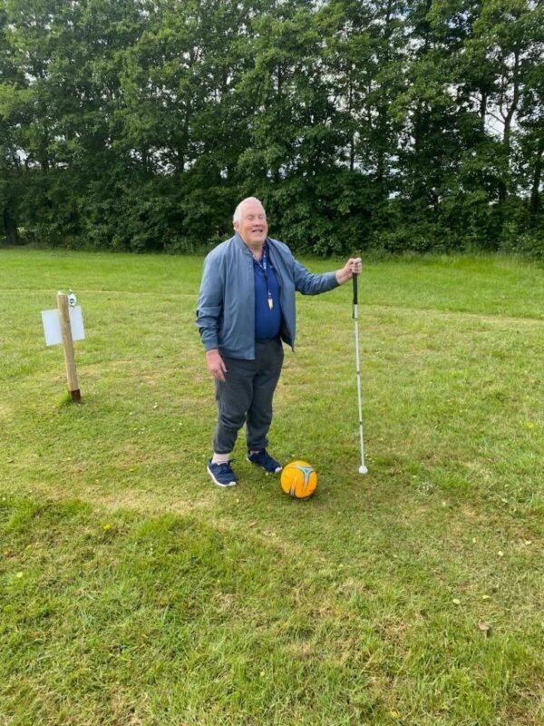 An older man supporting himself with a white cain and kicking a ball on a grass field