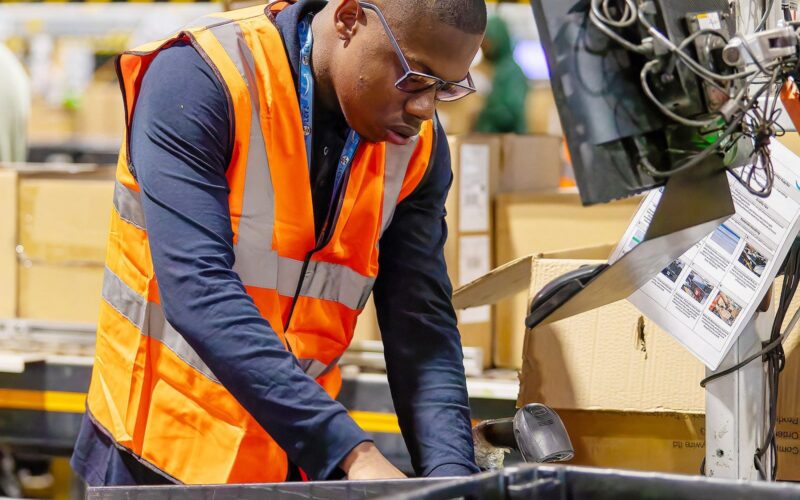 Young man with glasses working in a fulfillment centre, wearing an orange high visibility vest.