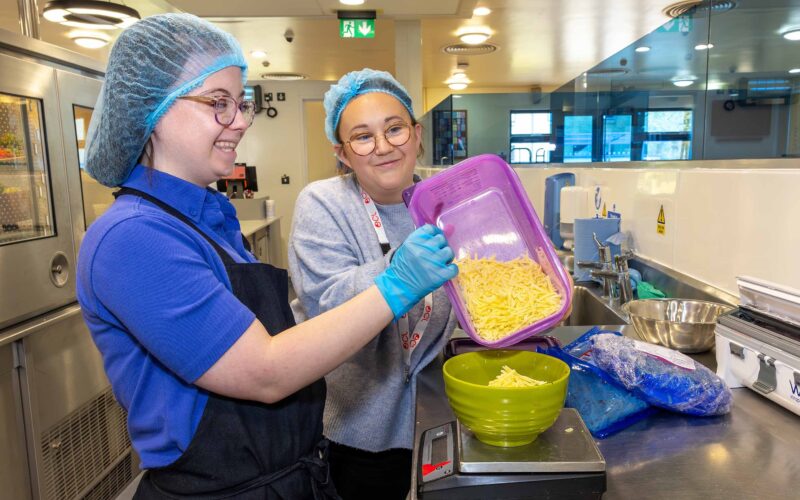 Young lady working in a canteen wearing a hairnet, glasses, apron and gloves.