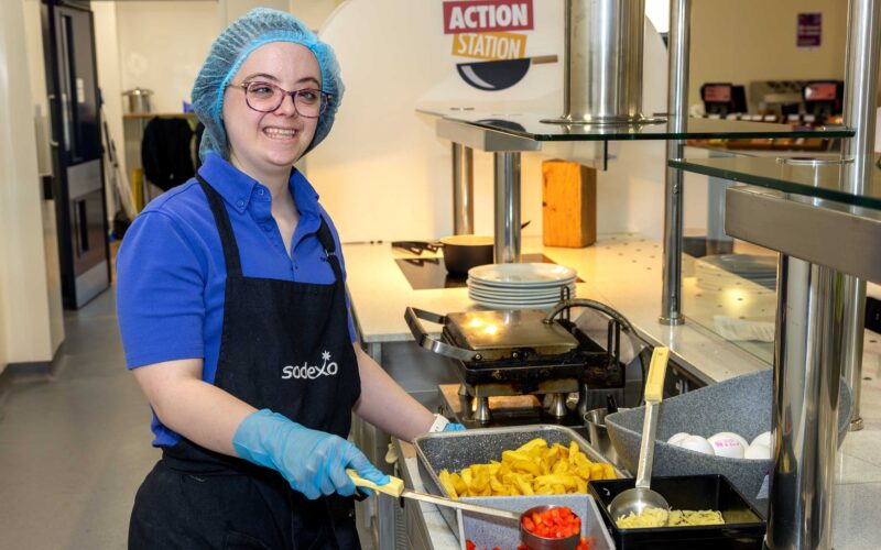 Young lady with a hairnet on and glasses, wearing an apron and gloves serving food in a canteen