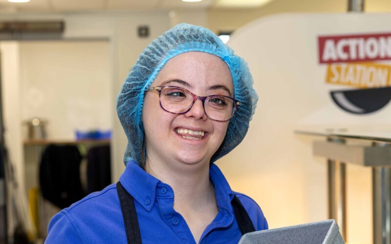 Young lady in a Sodexo Food Service Assistant uniform, with a hair net and glasses serving food in a canteen.