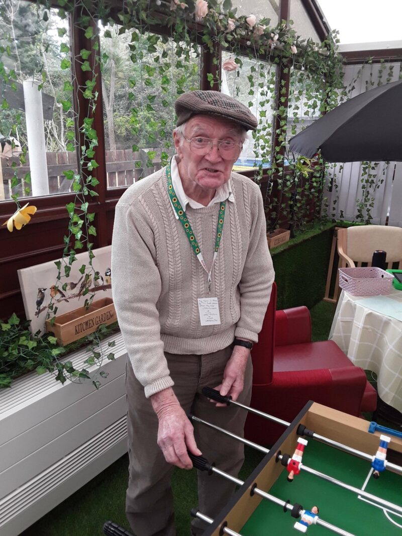 Elderly man standing up next to a football table smiling at the camera, getting ready to start playing.