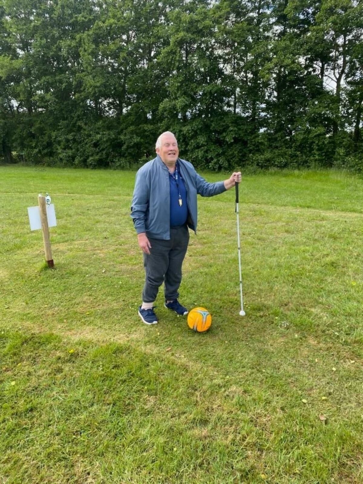 An older man supporting himself with a white cain and kicking a ball on a grass field