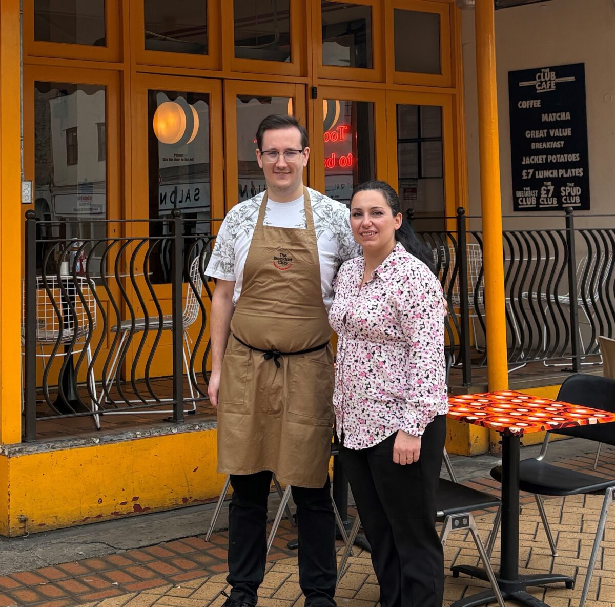 Young man with glasses and woman standing outside a cafe.