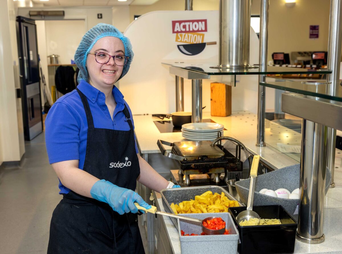 Young lady with a hairnet on and glasses, wearing an apron and gloves serving food in a canteen