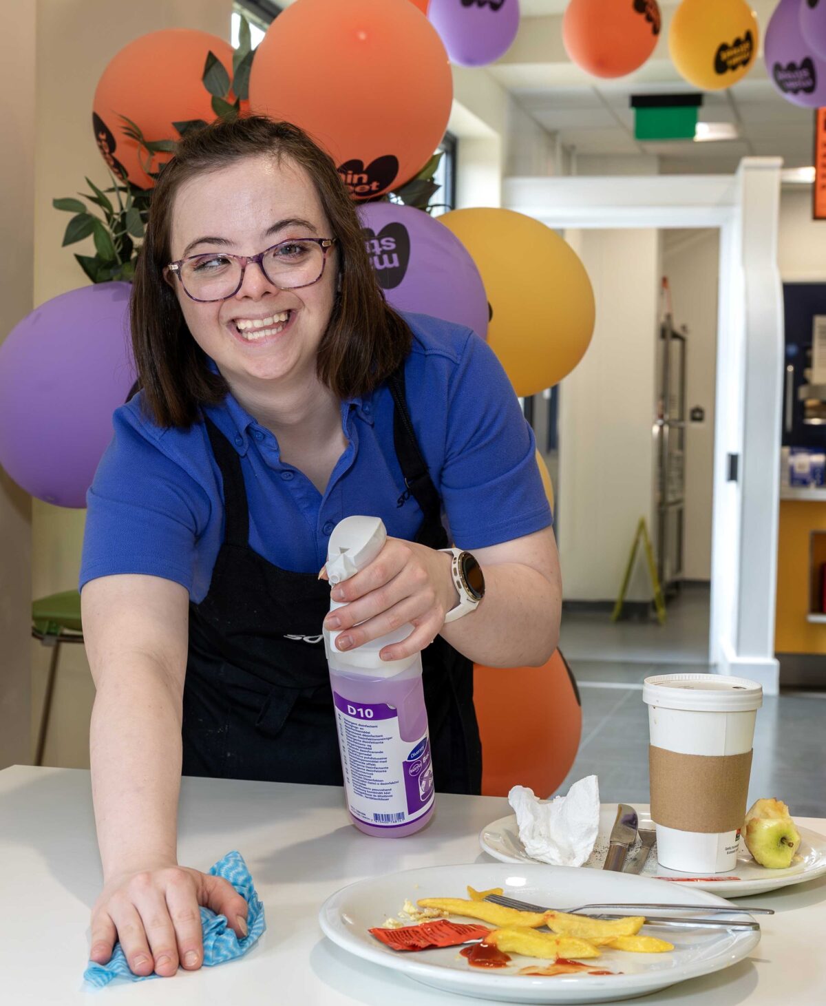 Young lady with glasses in Sodexo food service uniform clearing tables in a canteen.