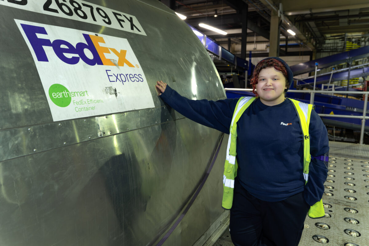 A young person in high-vis jacket standing next to a metal tank with FedEx logo on it