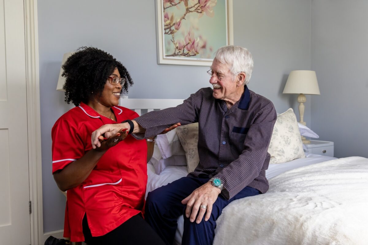 Woman in red tunic supports elder with rehabilitation near his bed-side