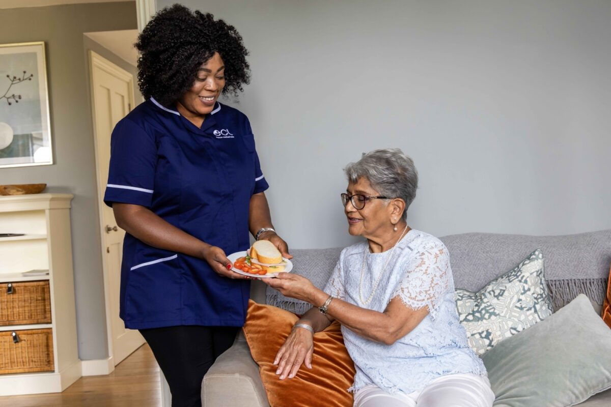 A woman on the left side of the photo is wearing a blue uniform with an ECL logo on it - she's handing an older woman on the right a plate with food