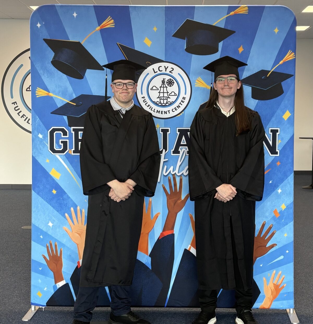 Two young men at their graduation, smiling, in caps and gowns with a 'congratulations' banner behind them.