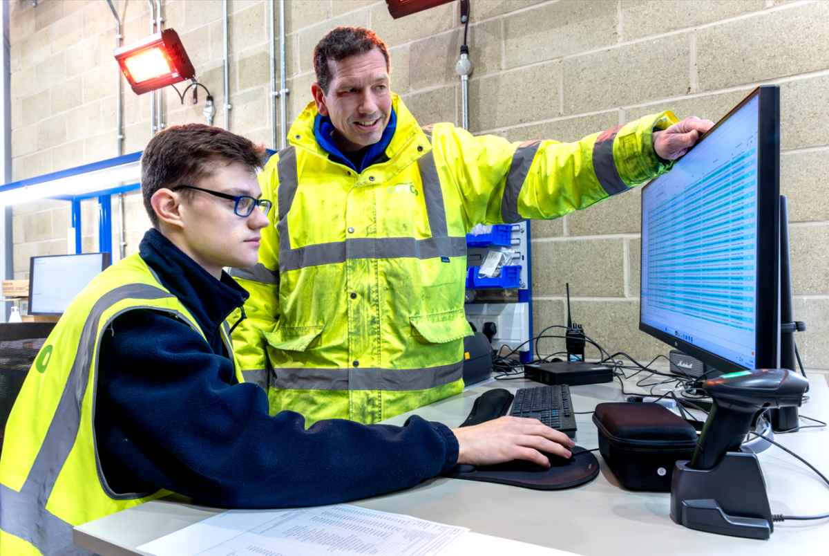 Two men in high-vis looking at a screen monitor, one of them is sitted, while the other is standing next to him and points at the monitor
