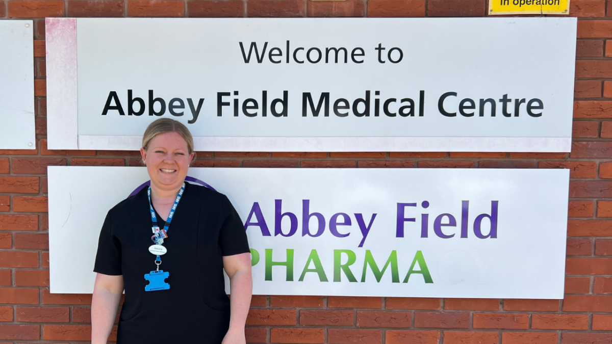 A blonde woman standing in front of a wall sign saying: 'Welcome to Abbey Field Medical Centre"