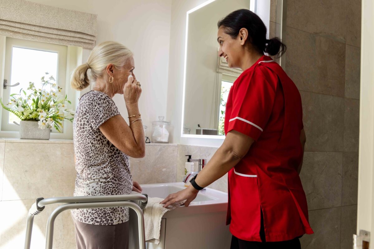 Older lady with carer at sink washing face.