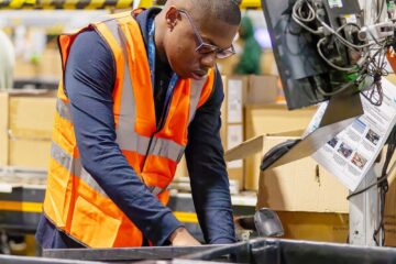 Young man with glasses working in a fulfillment centre, wearing an orange high visibility vest.