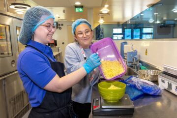 Young lady working in a canteen wearing a hairnet, glasses, apron and gloves.