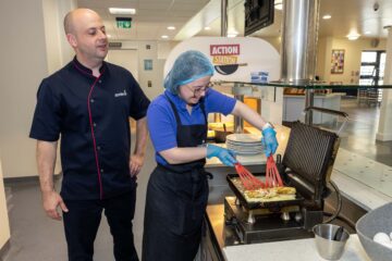 Man in chefs uniform with female food service assistant cooking an omelette in a canteen kitchen.