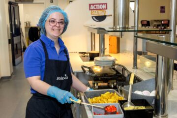 Young lady with a hairnet on and glasses, wearing an apron and gloves serving food in a canteen