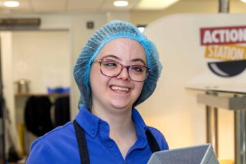 Young lady in a Sodexo Food Service Assistant uniform, with a hair net and glasses serving food in a canteen.