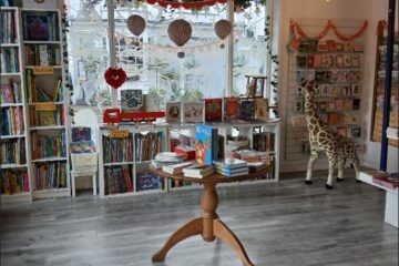 Internal photo of the new part of the shop showing the window display from the inside and a clear floor area with a small table display of books in the middle. There is a large giraffe in the right hand side of the shot.