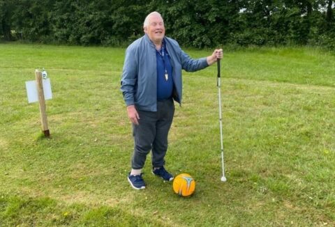An older man supporting himself with a white cain and kicking a ball on a grass field