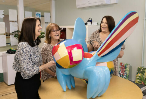 Three women standing around a light blue bee sculpture placed on the table