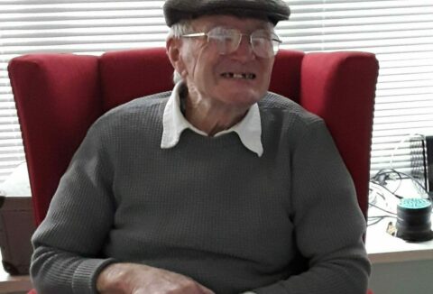 Elderly gentleman sitting down in a red armchair smiling at the camera.