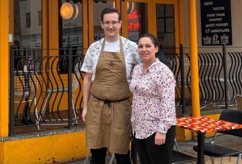 Young man with glasses and woman standing outside a cafe.