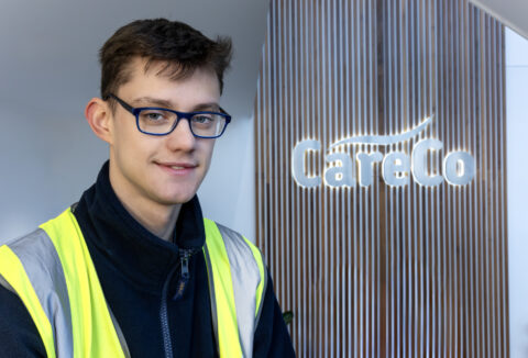 A young man in front of 'CareCo' sign wearing a high-ves