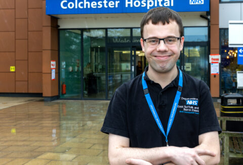 A young man standing in front of the Colchester Hospital looking at the camera and smiling