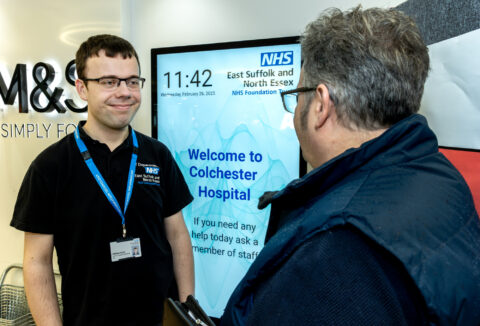 A young man talking with another man - in the background there's a sign saying 'Welcome to Colchester Hospital'