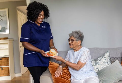 A woman on the left side of the photo is wearing a blue uniform with an ECL logo on it - she's handing an older woman on the right a plate with food