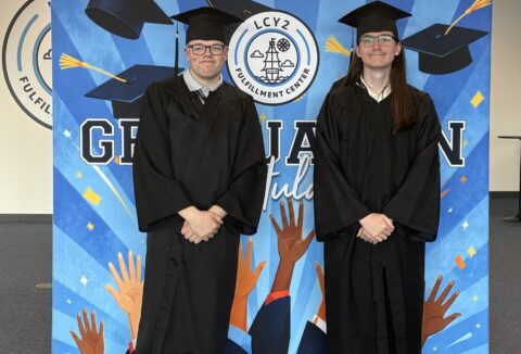Two young men at their graduation, smiling, in caps and gowns with a 'congratulations' banner behind them.