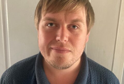 A blonde man facing the camera wearing blue polo shirt