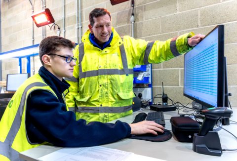 Two men in high-vis looking at a screen monitor, one of them is sitted, while the other is standing next to him and points at the monitor
