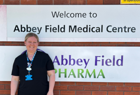 A blonde woman standing in front of a wall sign saying: 'Welcome to Abbey Field Medical Centre"