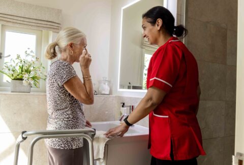 Older lady with carer at sink washing face.